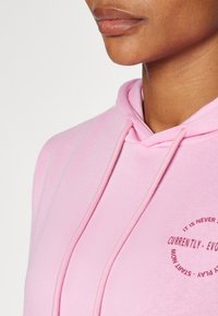 Hellrosa Kapuzenpullover mit Kordelzügen, der auf der Brust einen runden Grafikdruck mit braunem Text zeigt. Glattes Material mit lockerer Passform.
