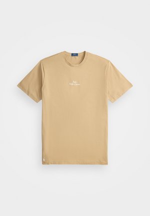 Beżowa koszulka z krótkim rękawem z okrągłym dekoltem, z białym logo "Polo Ralph Lauren" umieszczonym na środku klatki piersiowej oraz małym haftowanym emblematem w pobliżu dolnego brzegu.