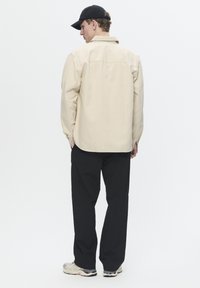Beige long-sleeve shirt with a relaxed fit and collar, paired with black pants. Model wears a black cap and light-colored sneakers.