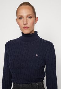Maglione a collo alto navy con motivo a treccia testurizzata, dotato di un piccolo logo ricamato sul petto e polsini a coste.