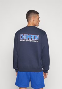 Sudadera azul marino con cuello redondo, logo en blanco y rojo "CHAMPION ATHLETICWEAR" en la parte posterior, puños y dobladillo acanalados.