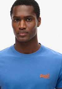 Camiseta de algodón azul con cuello redondo, que presenta un logotipo bordado naranja en el pecho. Textura suave, mangas cortas y un diseño ajustado.