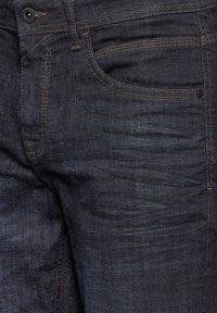 Mörka denimjeans med en närbild av tygstrukturen, med kontrasterande orangea sömmar och en detalj av framfickan.