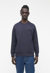 Tmavě námořnická mikina s vroubkovaným kulatým výstřihem a manžetami; na hrudi je vyražené logo. Hladká textura, volný střih, ve spojení s modrými džíny.