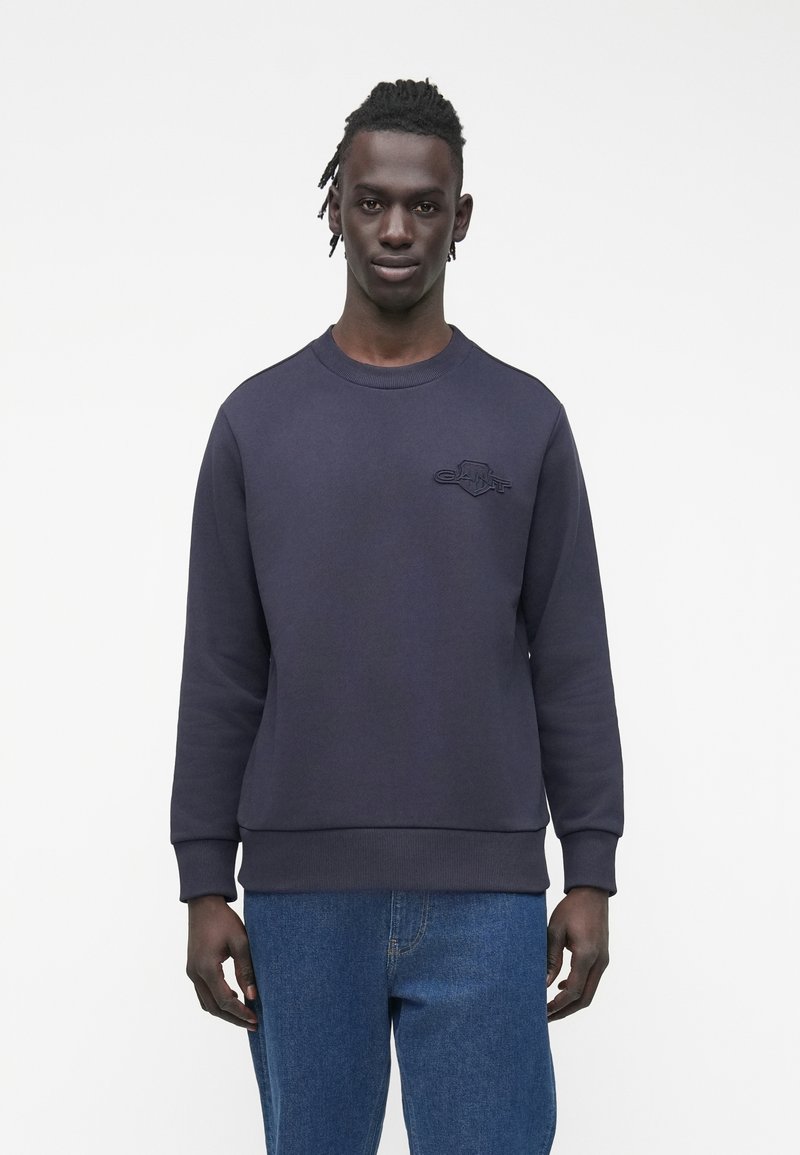 Tmavě námořnická mikina s vroubkovaným kulatým výstřihem a manžetami; na hrudi je vyražené logo. Hladká textura, volný střih, ve spojení s modrými džíny.