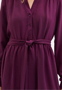 Långärmad klänning i djupt lila tyg med V-ringning, knappframsidan och ett knytbälte i midjan. Texturen verkar mjuk med en lätt lyster.