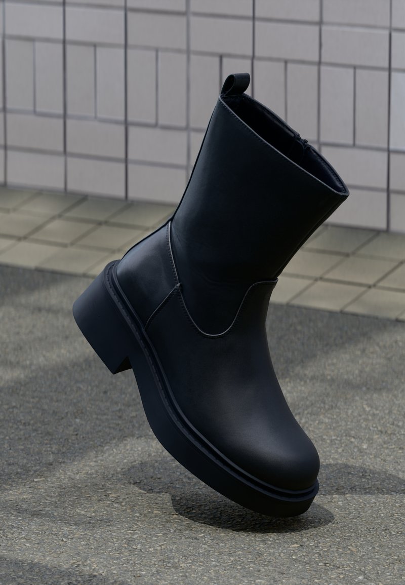 Μαύρη δερμάτινη μπότα μέχρι τη μέση της γάμπας με παχιά σόλα και τακούνι block σε πλακόστρωτο έδαφος μπροστά από φόντο με πλακάκια.