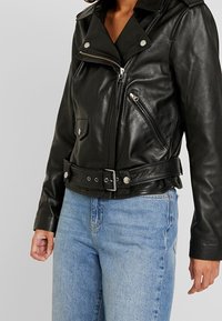 Czarna skórzana kurtka motocyklowa z kieszeniami zapinanymi na zamek, zatrzaskami i paskiem w talii. Gładka faktura, dopasowany fason i szeroki kołnierz.