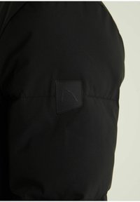 Manga de chaqueta negra acolchada con un parche cuadrado cosido que presenta un emblema minimalista de hoz en la parte superior del brazo sobre un fondo liso.