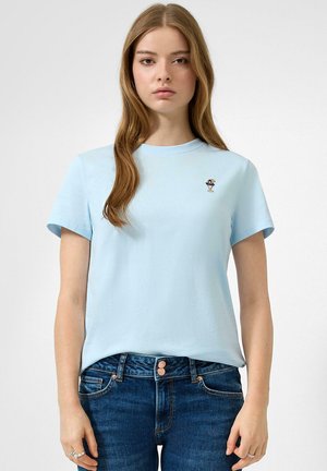 Junge Frau trägt ein hellblaues T-Shirt mit einer kleinen Stickerei eines Cocktailglases, kombiniert mit blauen Jeans, steht vor einem schlichten Hintergrund.