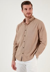 Camicia a maniche lunghe color marrone chiaro con colletto a bottoni, caratterizzata da una vestibilità relaxed e bottoni scuri, abbinata a pantaloni bianchi.