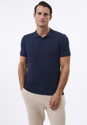 Camisa polo de manga corta en color azul marino, hecha de tejido de punto texturizado con un patrón de trenzas, con cuello y tres botones, combinada con pantalones de color claro.