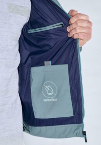 Wasserdichte Jacke mit marineblauem Innenfutter, ausgestattet mit einer hellgrauen Tasche, auf der ein wasserdichtes Symbol aufgedruckt ist, sowie einer inneren Reißverschlusstasche.