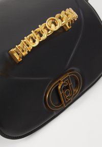 Borsa a mano in pelle nera con manico a catena dorata, caratterizzata da scritte stilizzate. I dettagli in hardware dorato con logo sulla parte frontale aggiungono contrasto e particolare attenzione.