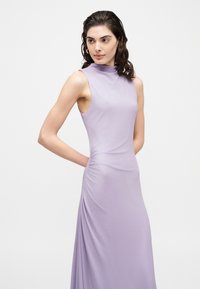 Femme aux cheveux foncés portant une robe ajustée sans manches, de couleur lavande et à col haut, debout les mains derrière le dos, regardant de côté.