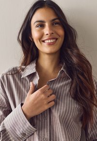 Jeune femme souriante aux longs cheveux foncés portant une chemise grise rayée à boutons, tenant le col avec sa main droite.