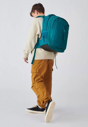 Ragazzo con maglione beige, pantaloni cargo marroni, sneakers nere, che porta uno zaino grande turchese, camminando su sfondo uniforme.