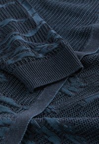 Mørk marineblåt strikket stof, der viser detaljerede teksturerede mønstre og foldet ribstrikket manchet på et langærmet beklædningsstykke.