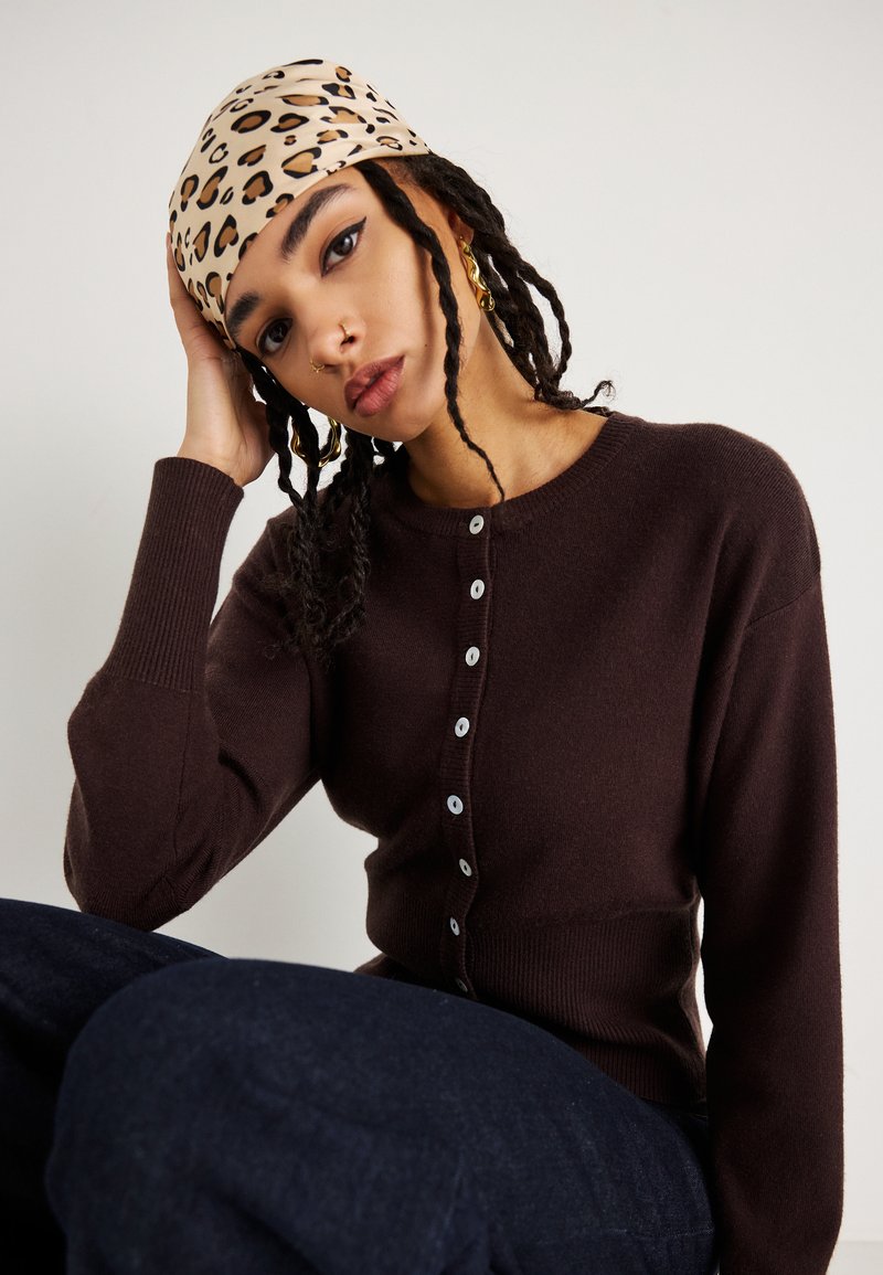 Cardigã castanho tricotado com decote redondo, apresentando seis botões e punhos canelados. O modelo usa um lenço de cabeça com padrão de leopardo.