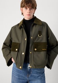 Olijfgroene jas met kraag, gouden hardware en corduroy zakken. Voorzien van een ritssluiting en een kenmerkende ringdetail op de schouder.