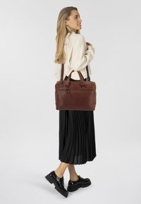 Borsa a mano in pelle marrone con manici doppi e tracolla removibile. Presenta una tasca con zip e una texture liscia. Il modello indossa una gonna nera plissettata.