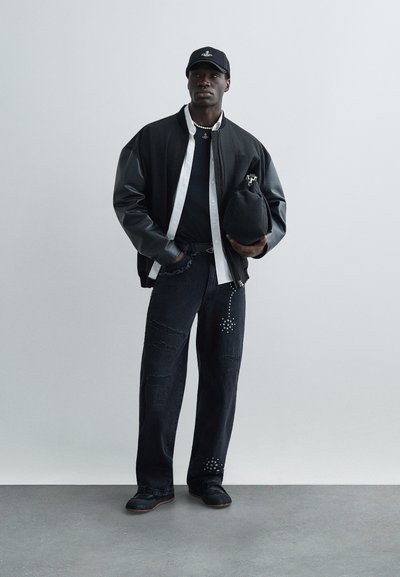 Μαύρο bomber μπουφάν με δερμάτινα μανίκια, λευκό πουκάμισο, μαύρο T-shirt, σκισμένο μαύρο τζιν με διακοσμητικά στοιχεία και μαύρα παπούτσια. Κρατάει μια μαύρη τσάντα.