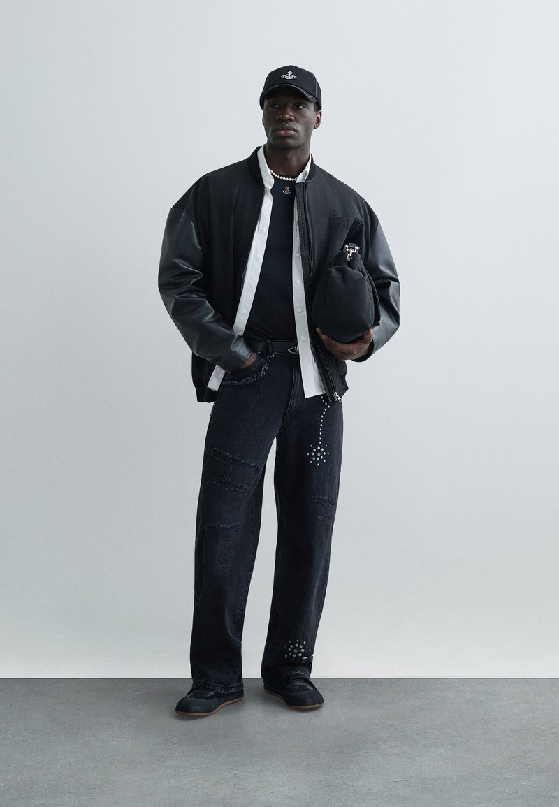 Black bomber jacket with leather sleeves, white shirt, black T-shirt, distressed black jeans with embellishments, and black shoes. Holding a black bag.