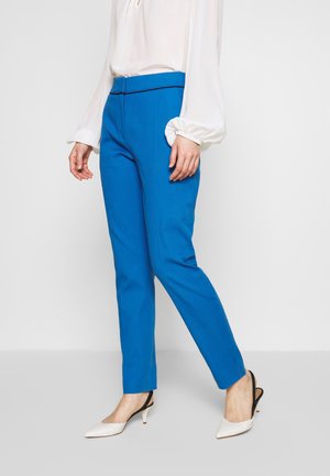 Kobieta ubrana w jaskrawo niebieskie dopasowane spodnie, białą bluzkę z długimi bufkami oraz białe sandały na obcasie z czarnymi paskami.