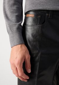 Černé kožené kalhoty s hladkou texturou, se hnědým logem Calvin Klein a zlatými kovovými doplňky. Spárované se šedým svetrem na rukávech.