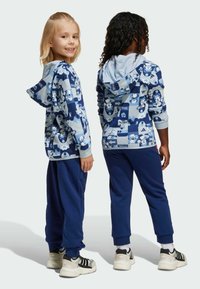 Niebieska bluza z kapturem z nadrukami w stylu kreskówkowym, jasnoniebieskie wykończenie, oraz niebieskie dresy. Miękka tkanina, luźny krój, ściągacze przy mankietach. Sneakersy z czarnymi akcentami.