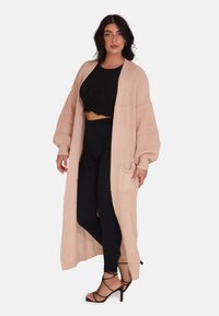 Lång, öppen cardigan i ljusbeige stickad kvalitet, med ribbat mönster och överdimensionerade ärmar, bärs över en svart crop top och leggings.