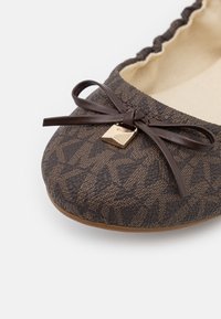 Chaussure plate décorative marron avec surface texturée, nœud accentué et détail en métal doré. Doublure intérieure douce. Conception à bout arrondi.