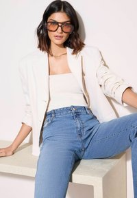 Blazer blanco sobre una blusa cremosa, jeans azules de cintura alta. Modelo con gafas de sol marrones de gran tamaño. Superficie texturada y fondo neutro.