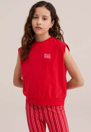 Tüdruk pikkade pruunide juustega kannab punast varrukateta toppi tekstiga „FLOWER“ ning punase-valgete triibuliste pükstega, seisab käed selja taga.