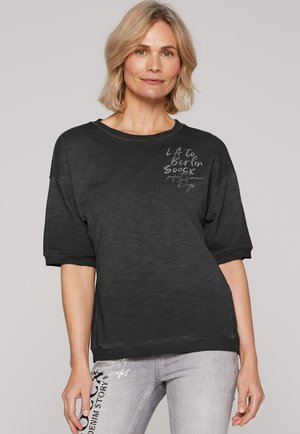 Schwarzer, übergroßer Sweatshirt mit kurzen Ärmeln, verziert mit weißem Text auf der linken Seite. Getragen mit hellgrauen Jeans, die bedruckte Details zeigen.