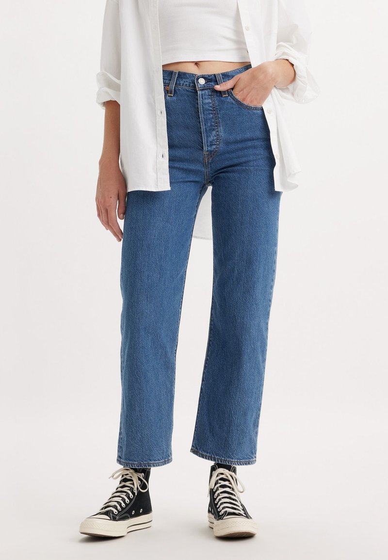 Jean en denim bleu taille haute à coupe droite, avec une fermeture à boutons, des coutures discrètes, portés avec des baskets montantes noires.