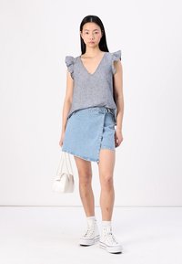 Grå V-hals topp med volang-sleeves kombinert med et lys blått denimskjørt. Modellen holder en hvit håndveske og har på seg hvite høye sneakers.