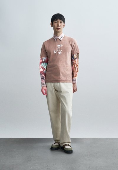 Καφέ κοντομάνικη μπλούζα με λευκό γραφικό, φορεμένη πάνω από πολύχρωμη μακρυμάνικη μπλούζα. Μπεζ παντελόνι φαρδύ και μαύρα παπούτσια με λευκές λεπτομέρειες.