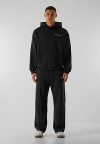 Hoodie noir avec poche avant et logo en texte sur la poitrine, associé à un pantalon de survêtement noir assorti. Matériau en coton, coupe décontractée, design minimaliste.