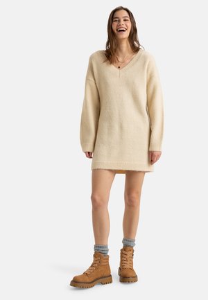 Beige neulemekko, jossa on V-pääntie, pitkät hihat, ribattua helmaa ja teksturoitu kangas; yhdistettynä vaaleanruskeisiin nauhakenkiin ja harmaisiin sukkiin.