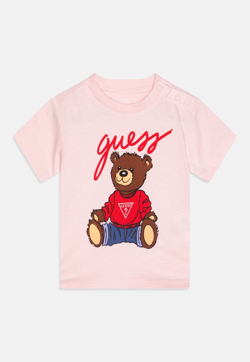 T-shirt rose à manches courtes présentant un graphisme d'un ours dans un pull rouge et un pantalon en denim, avec "guess" en lettres rouges en gras au-dessus.