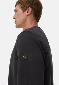 Zwarte sweatshirt met een ronde hals en zijkantdetail. Heeft een gestructureerde zwarte patch met een oranje logo op de linkerschouder.