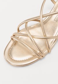 Sandale en métal doré avec des lanières croisées, un bout carré et une texture en cuir lisse. Mise en valeur par des coutures détaillées.