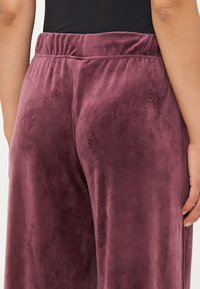 Pantaloni din catifea într-o nuanță bogată de prună, cu o textură moale, având un elastic la talie și un finisaj neted, prezentați din spate.