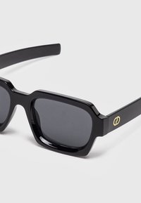 Gafas de sol rectangulares negras con montura gruesa y lentes oscuras, con un pequeño logo dorado en la patilla.