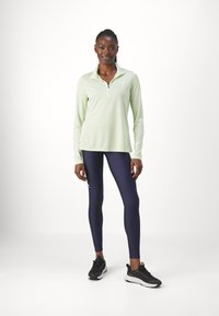 Lysgrøn langærmet atletisk top med halv-zip, parret med tætsiddende marineblå leggings og sorte løbesko. Et logo vises på toppen.
