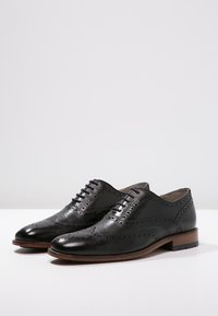 Chaussures noires en cuir à lacets avec des détails brogue, présentant une finition lisse, un bout rond et une semelle en cuir marron.