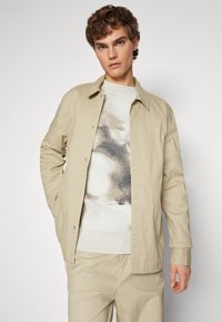 Chaqueta ligera color beige con cierre de botones, un bolsillo en el pecho, llevada sobre un suéter texturizado con patrón crema y beige. Acabado de tela suave.