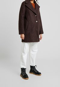 Brun, texturerad ullkappa med slagkrage, två knappar och sidofickor, parat med vita jeans och svarta snörkängor.