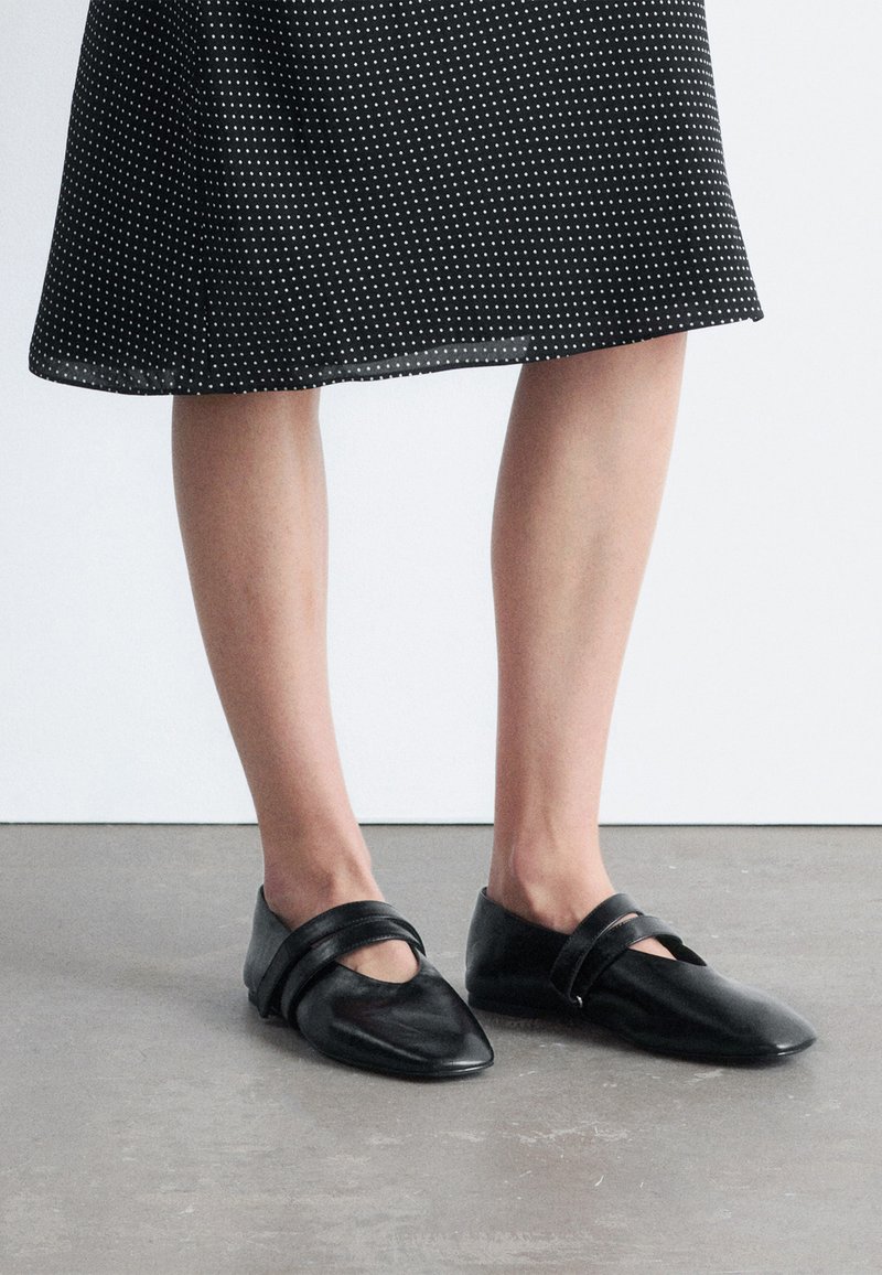 Bas des jambes portant une jupe noire à pois blancs et des chaussures plates Mary Jane en cuir noir sur un sol gris.